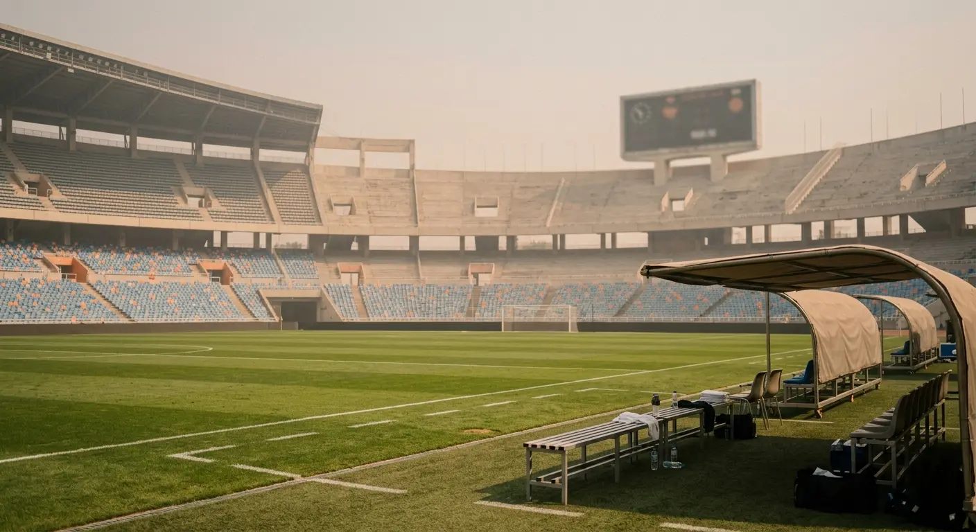 Empty open-air stadium under hazy heat-dome sky, scoreboard softly out of focus, single shaded technical area in foreground, afternoon harsh light, warm desaturated palette